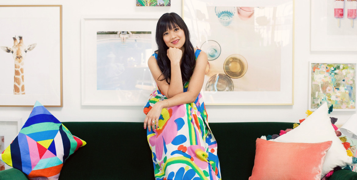 Joy Cho sits on the back of her couch at home in brightly patterned dress, photo