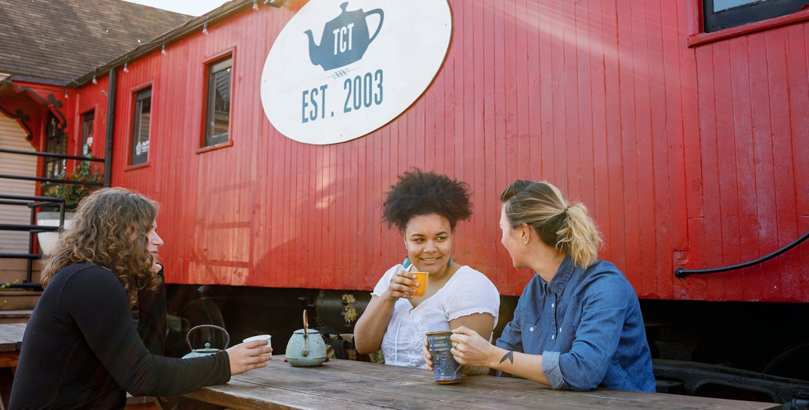 three women seated outdoors at picnic table share a pot of tea beside the red caboose that houses Tea Chai Té in Portland, Oregon, picture