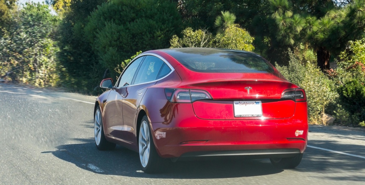 Red Tesla Model 3 driving with carpool stickers on the bumper.