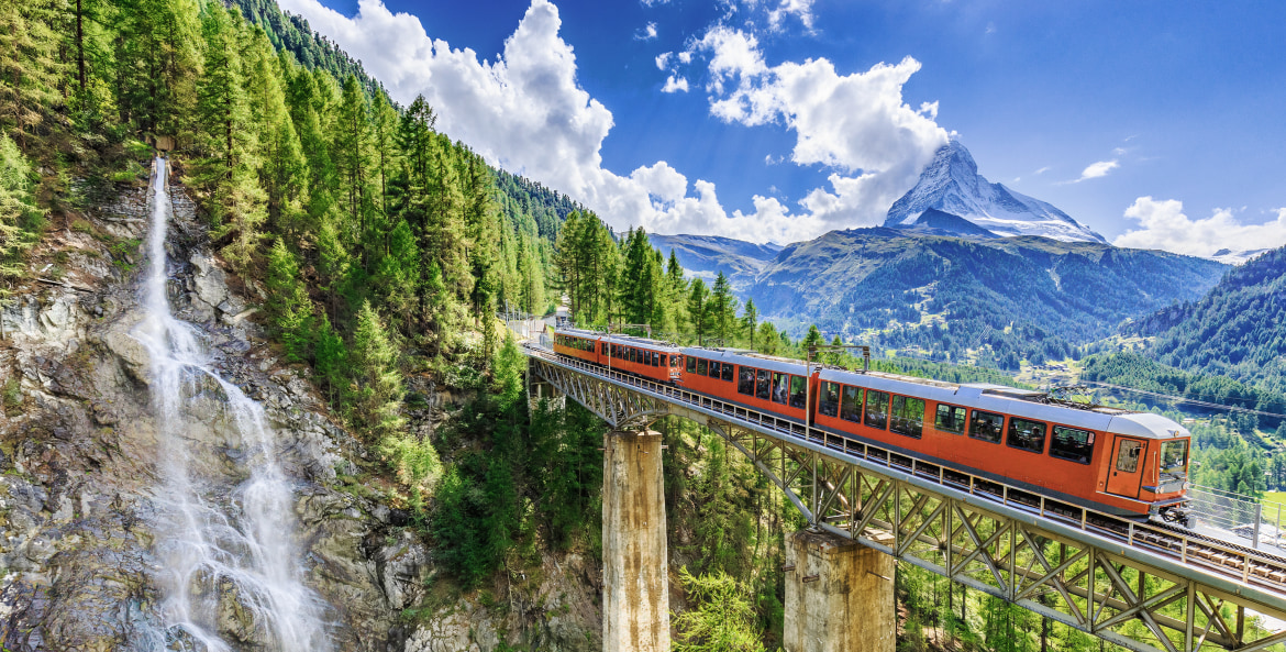 A train crosses a bridge by a waterfall in the Swiss Alps, image