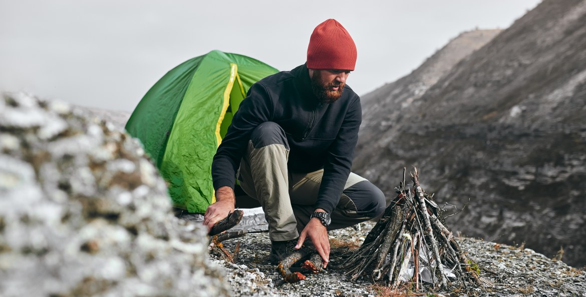 A hiker builds a campfire outside a tent on a mountainside, image