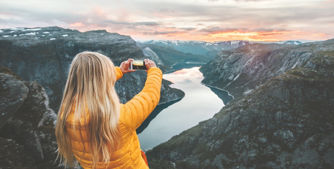 A woman in a yellow jacket takes a picture of a fjord in Norway at sunrise or sunset