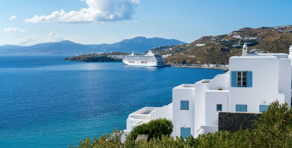 view of a cruise ship docked beyond a hotel and across the blue waters in Mykonos, Greece