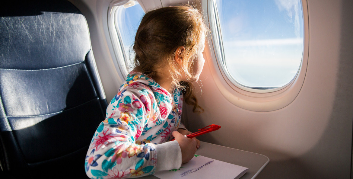 A little girl takes a break from coloring to look out the airplane window.