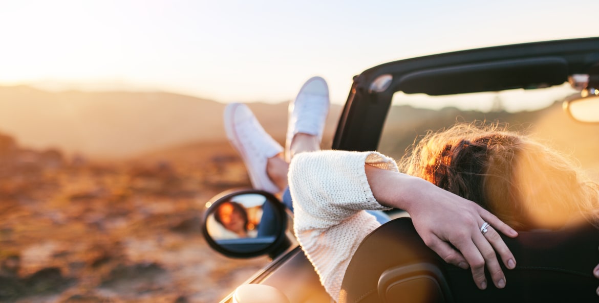 A woman sits with her feet out the window of a convertible at sunset.