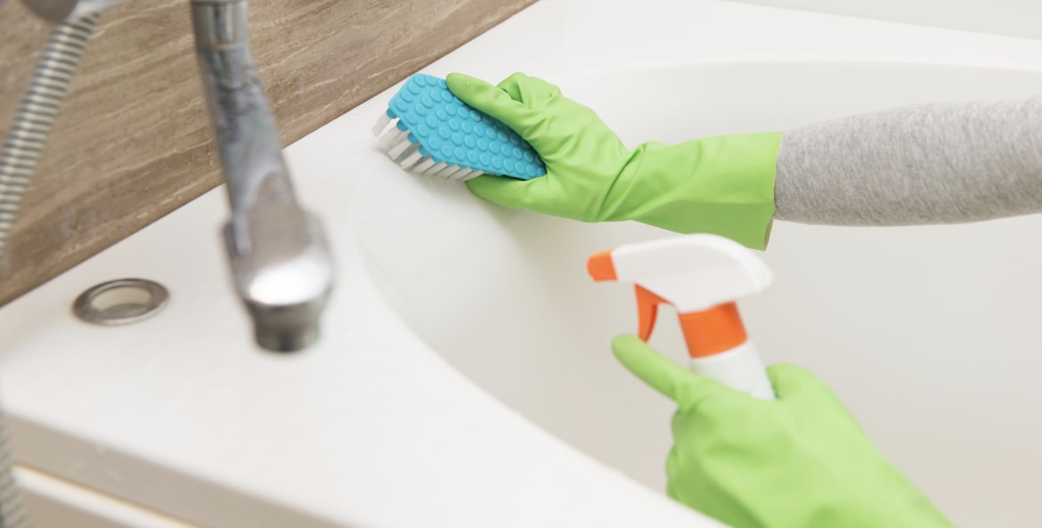 a gloved hand scrubbing a bathtub with a sponge and spray bottle
