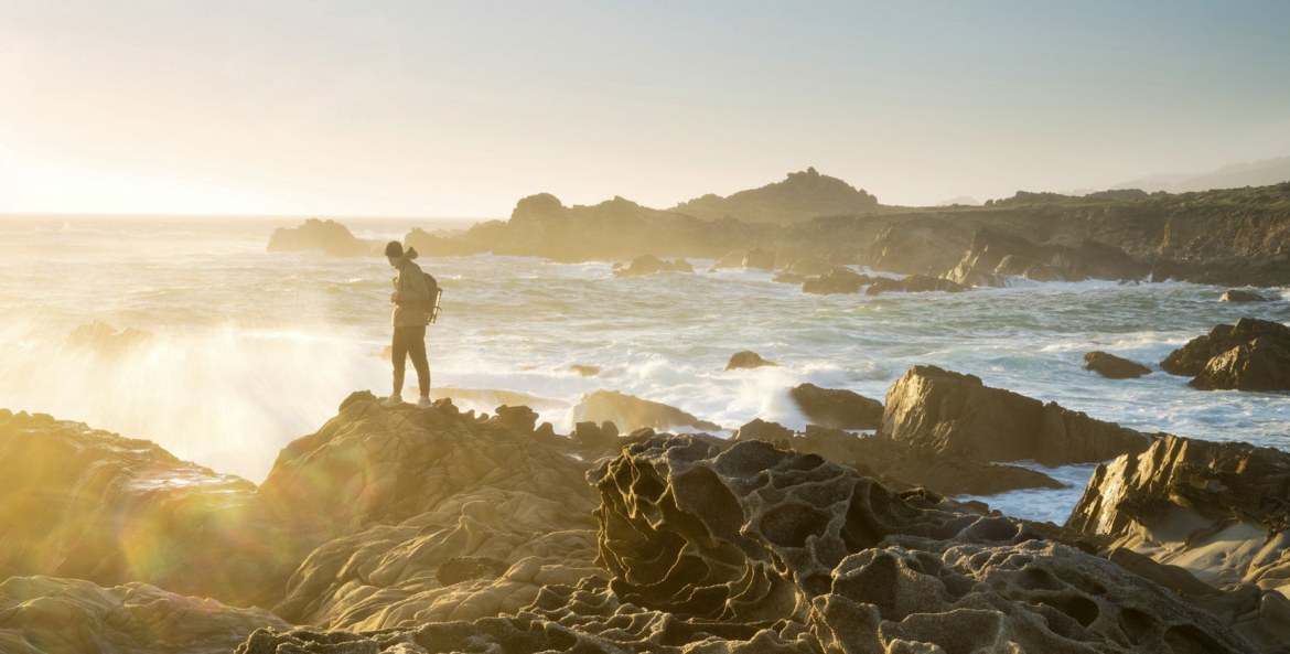 Hiker dressed warmly and standing on the honeycombed rocks known as tafoni looks out to sea at sunset at Salt Point State Park on the Sonoma Coast in California