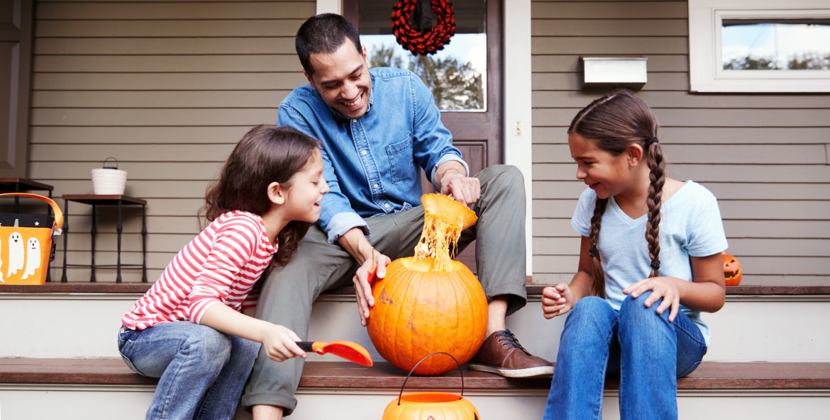 A dad carves a pumpkin with his daughters on their porch.
