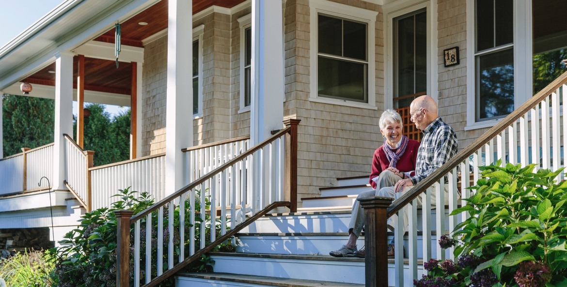 elderly couple sits smiling and laughing on the front steps leading to their home