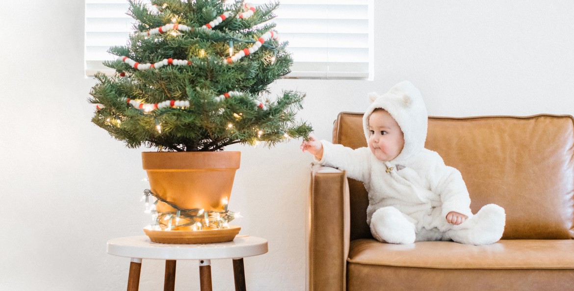 baby wearing romper seated on sofa reaches over to touch small potted Christmas tree placed on stool