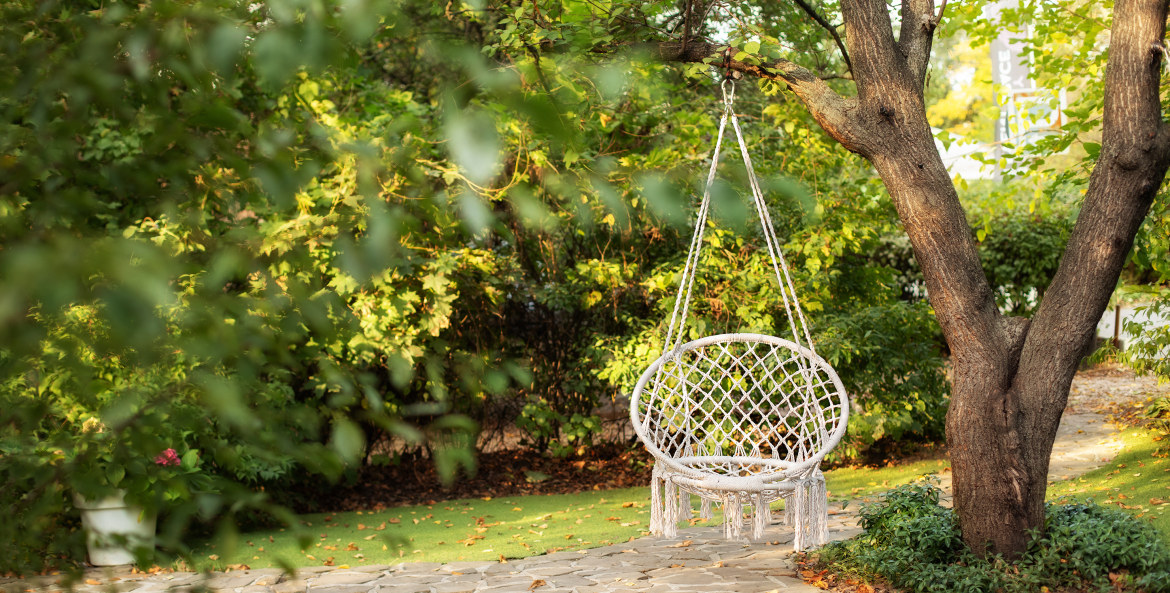 A white wicker chair hangs from a tree in a backyard.