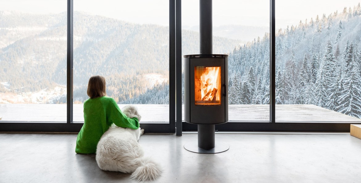 A woman and her dog sit in front of a wood-burning stove and look out the window at a snow-covered landscape.