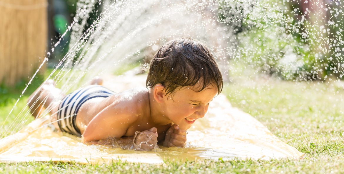 A kid lays in the water on a slip and slide on a hot day.