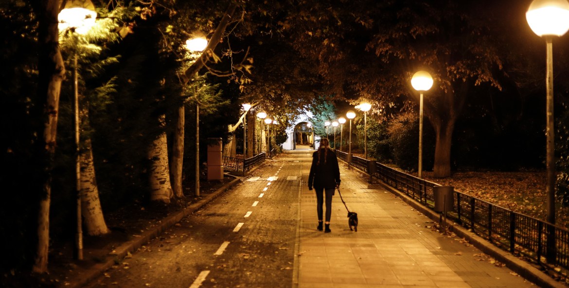 A woman walks her dog on a well-lit path through a park at night.