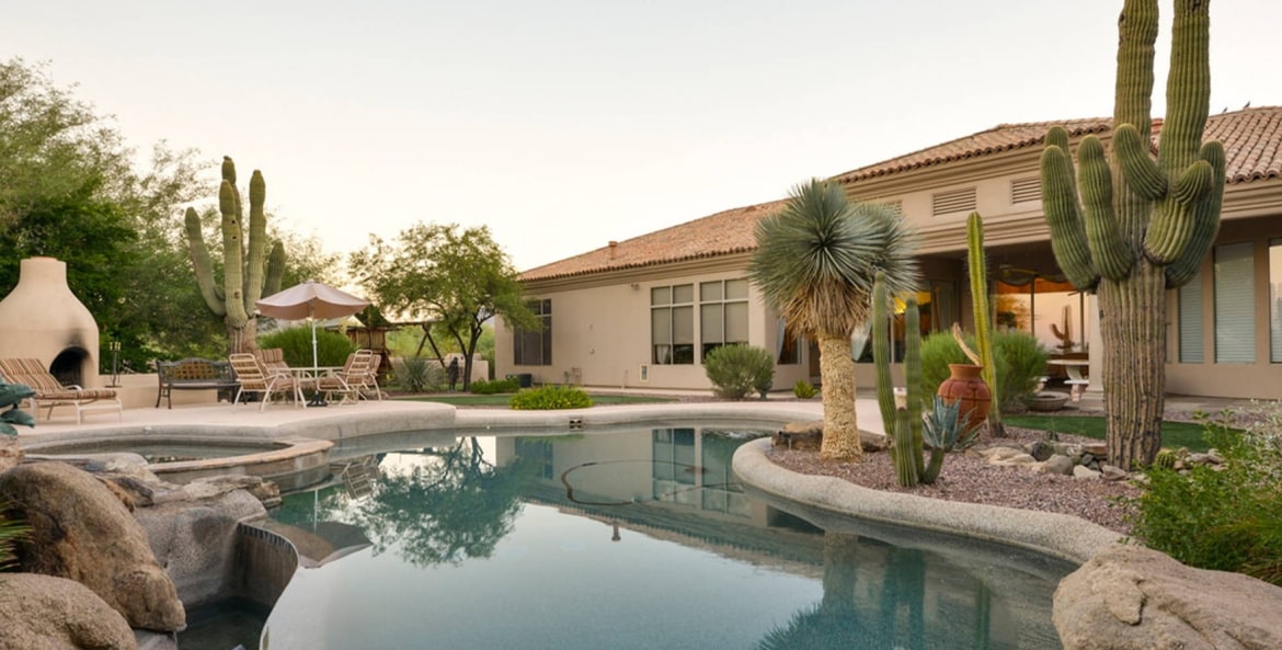 The backyard of a southwestern style home with a pool as the focal point. The pool is surrounded by a variety of cactus, rock scaping, a fireplace, and the back of the home can be seen.