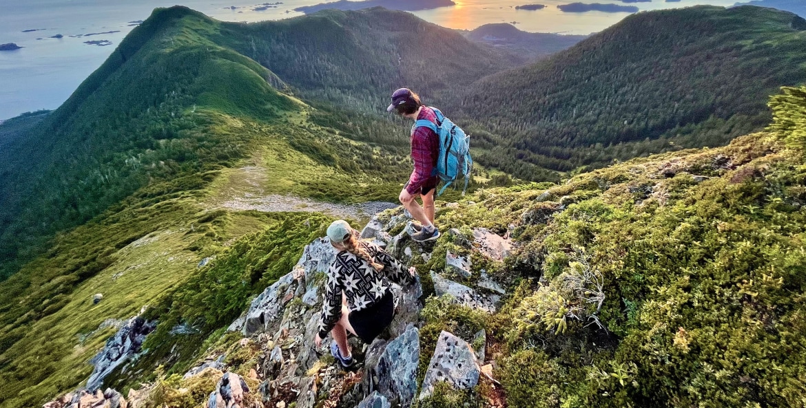 Two hikers scramble on rocks along trail overlooking Sitka sound at sunset. 