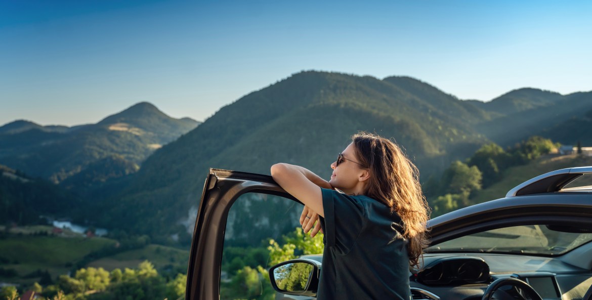 A woman stands by her open car door and looks out a mountain view.