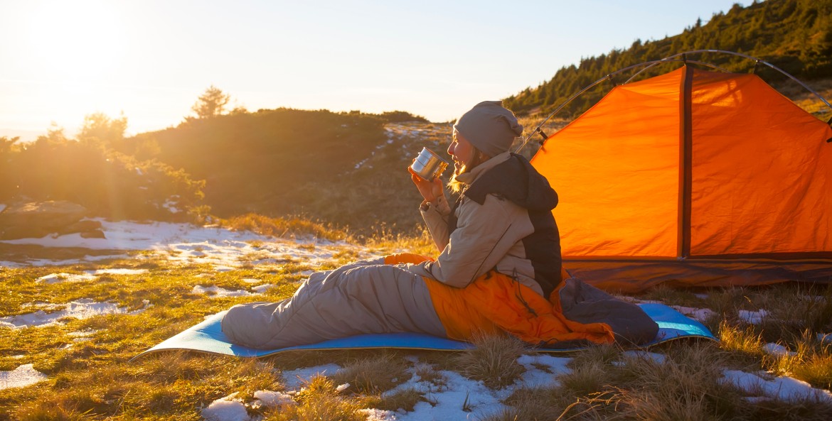 A camper sits on a sleeping pad and sleeping bag on frozen ground while drinking a coffee.