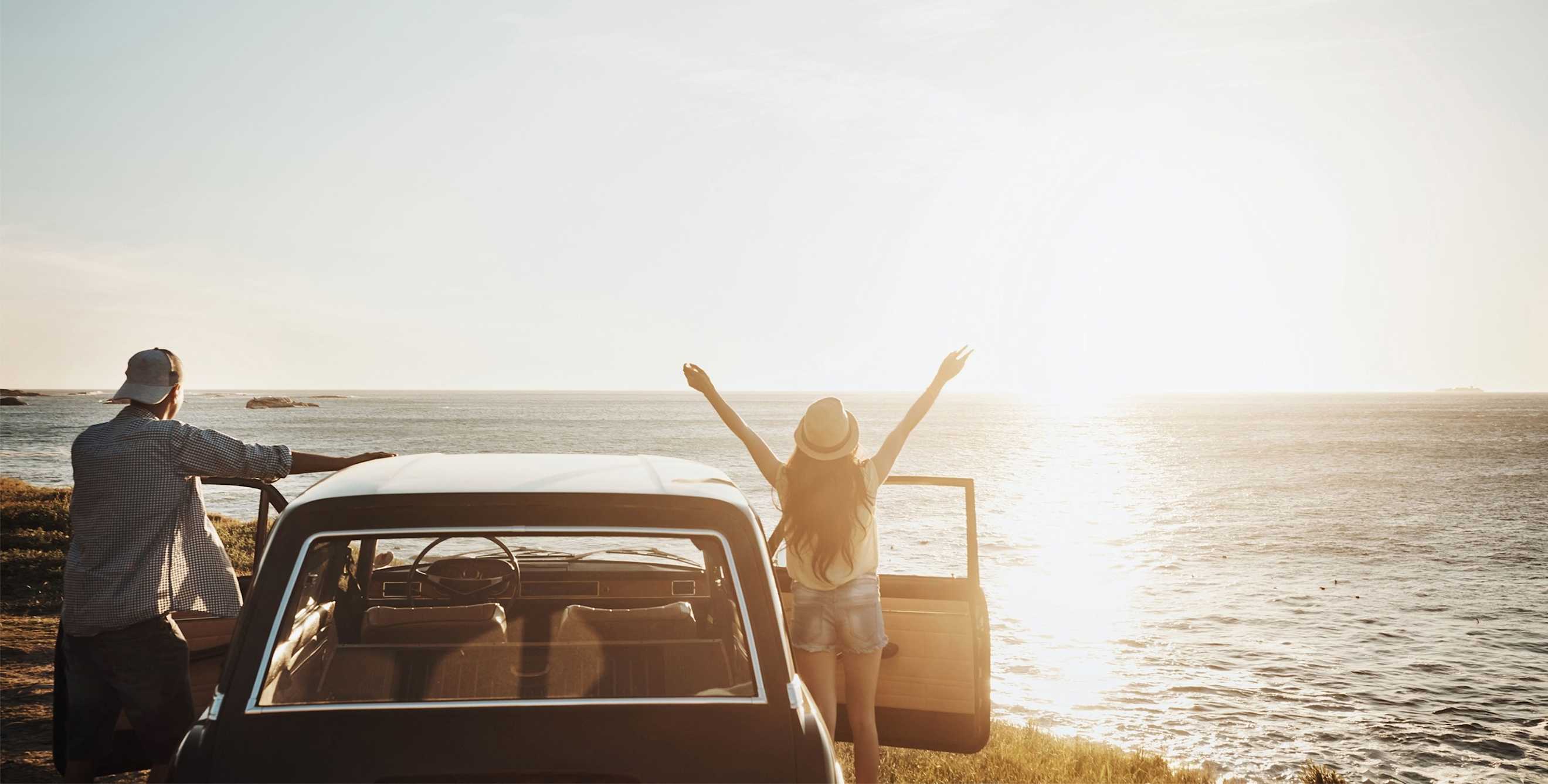 A couple on a road trip stand outside their car looking at the sun setting over the ocean