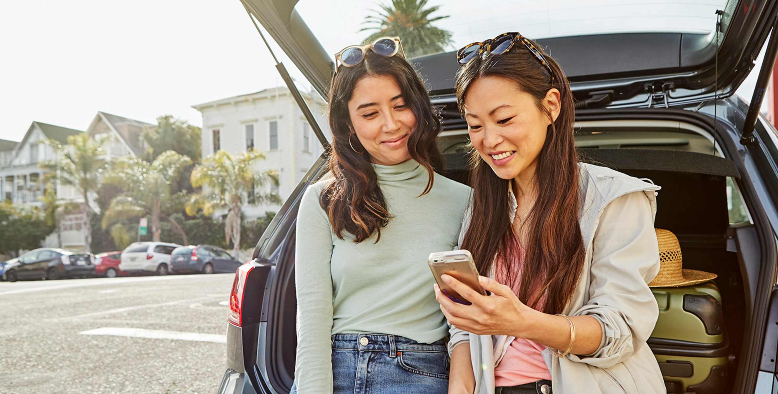 two women with smartphone seated on open SUV hatchback plan road trip