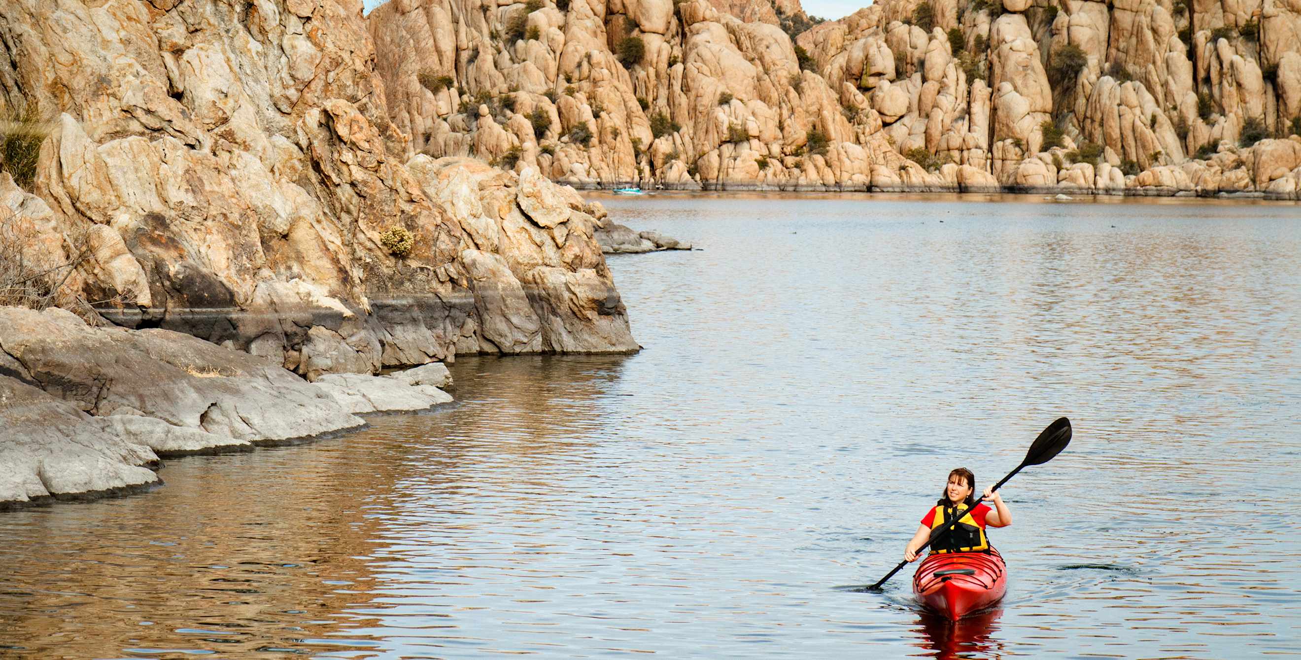 A woman paddles a kayak on Watson Lake in Prescott, Arizona with rocky walls behind her