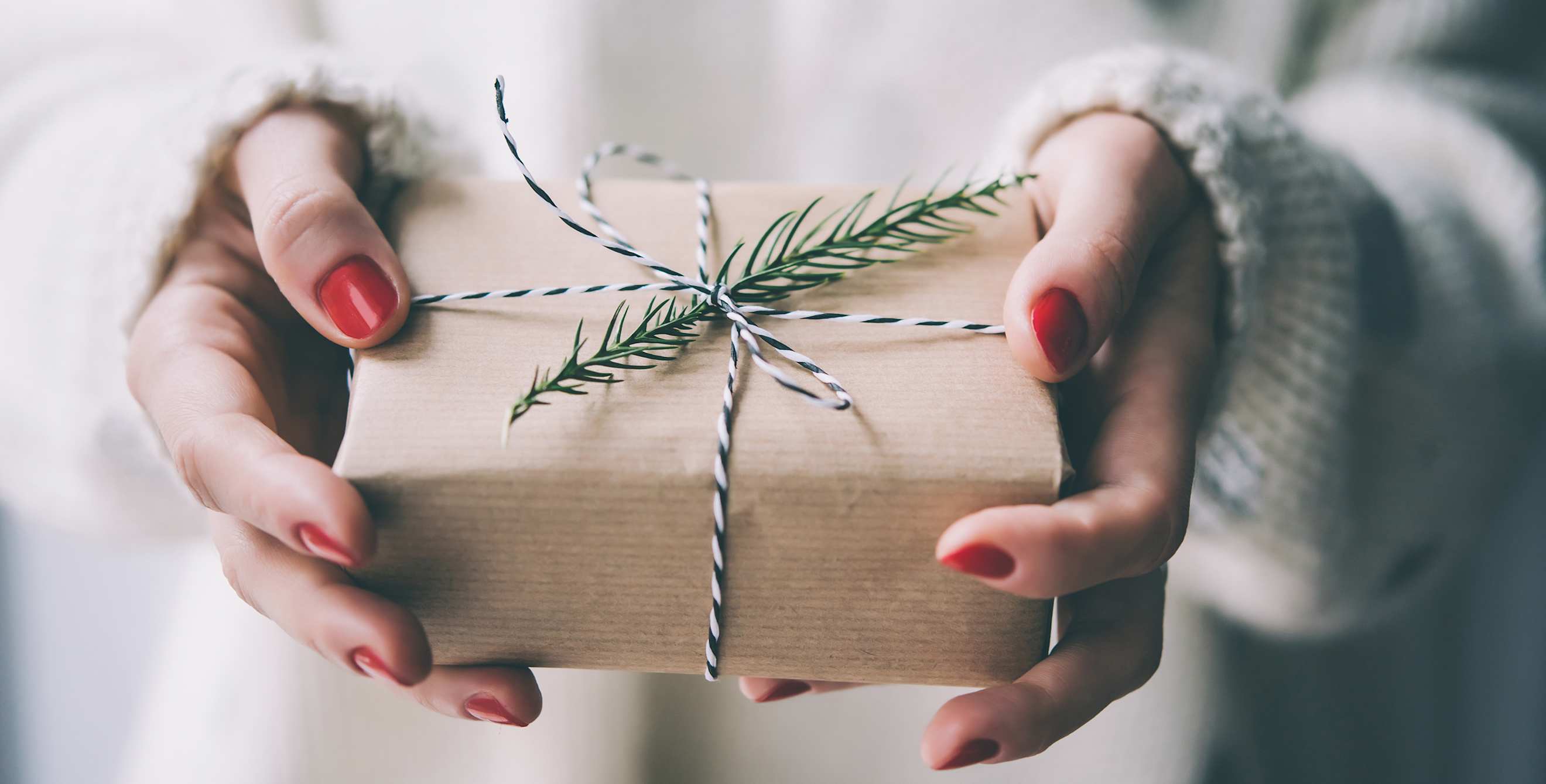 A woman in a polka dot sweater holds a sustainably wrapped modern present with twine and a sprig of evergreen branch, image