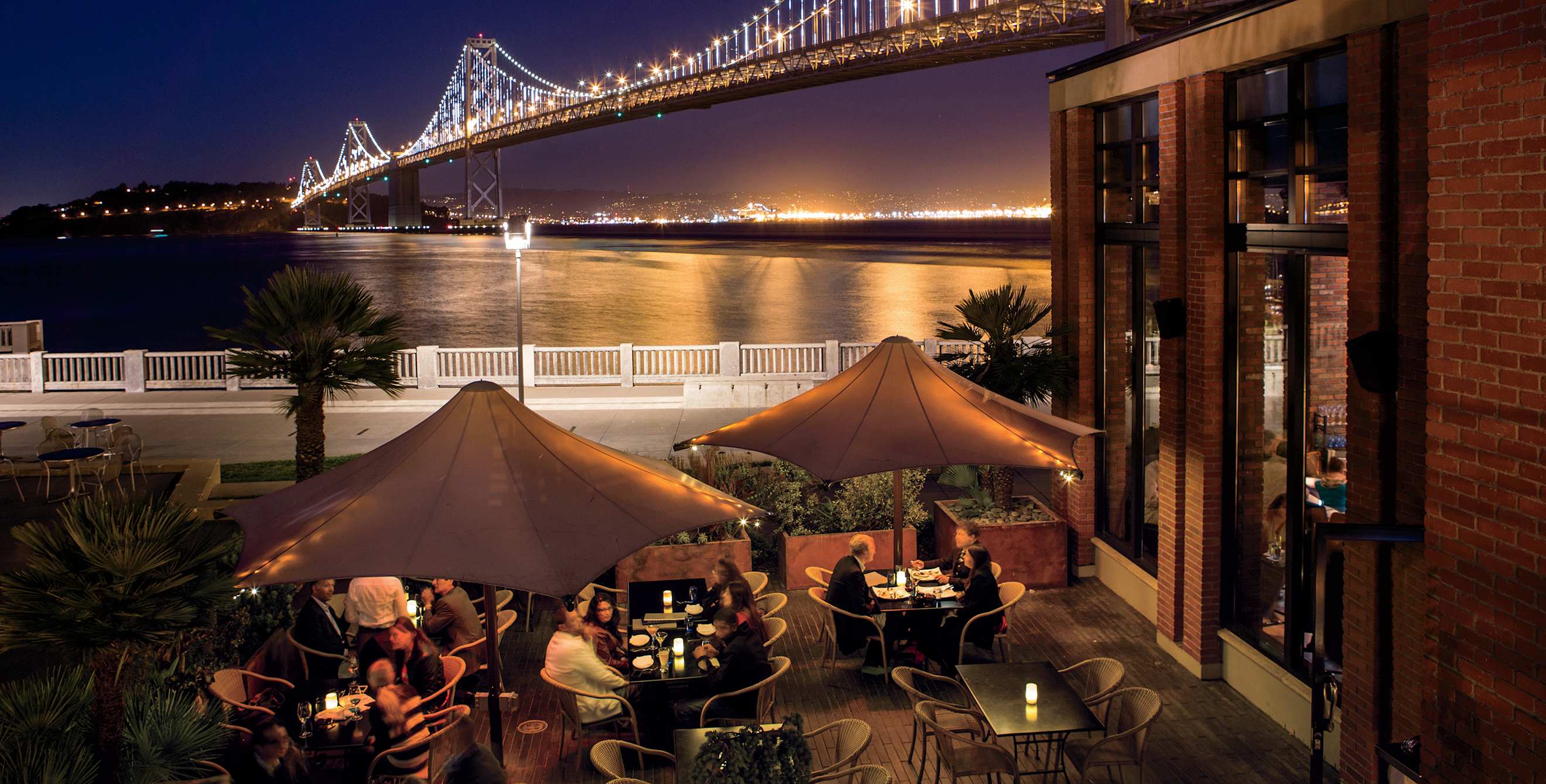 Waterbar diners seated outdoors under umbrellas at twilight along the Embarcadero next to the Bay Bridge in San Francisco
