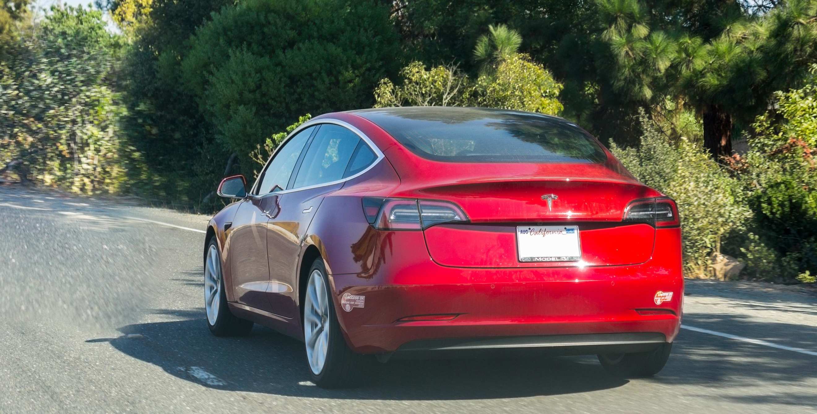 Red Tesla Model 3 driving with carpool stickers on the bumper.