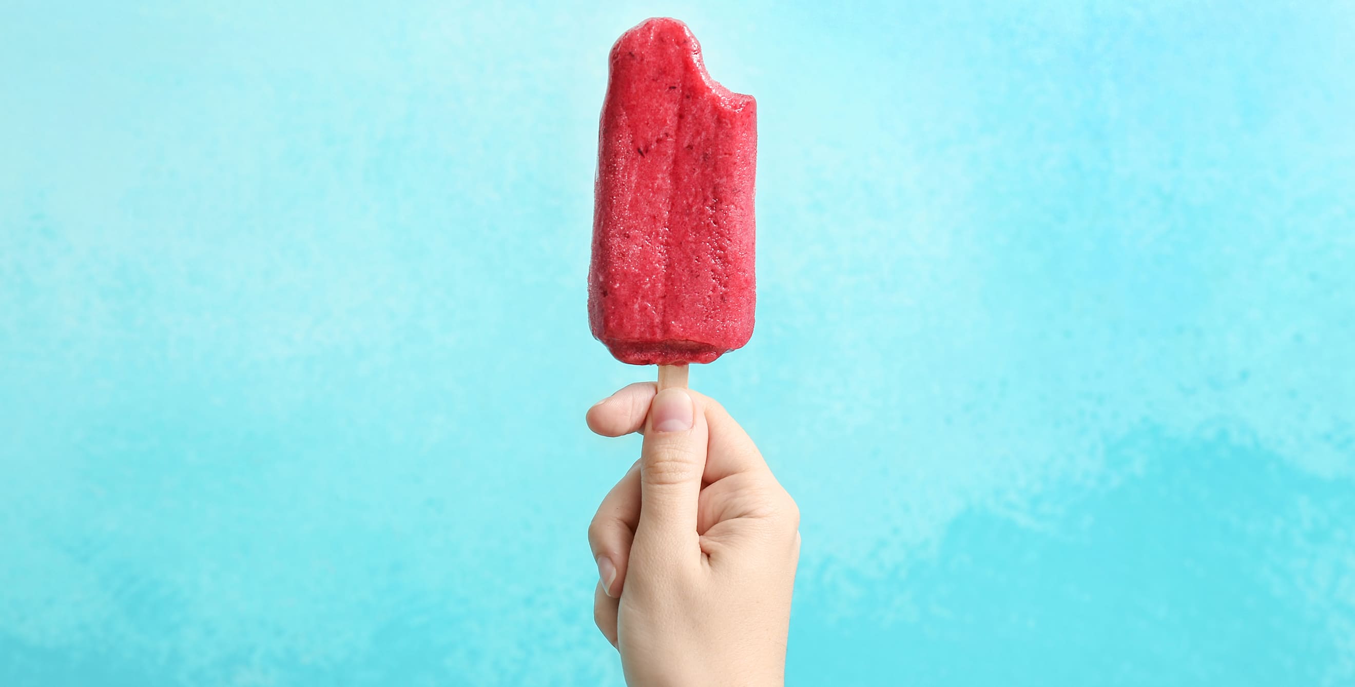 A hand holds a popsicle with a bite out of it against a blue background, photo