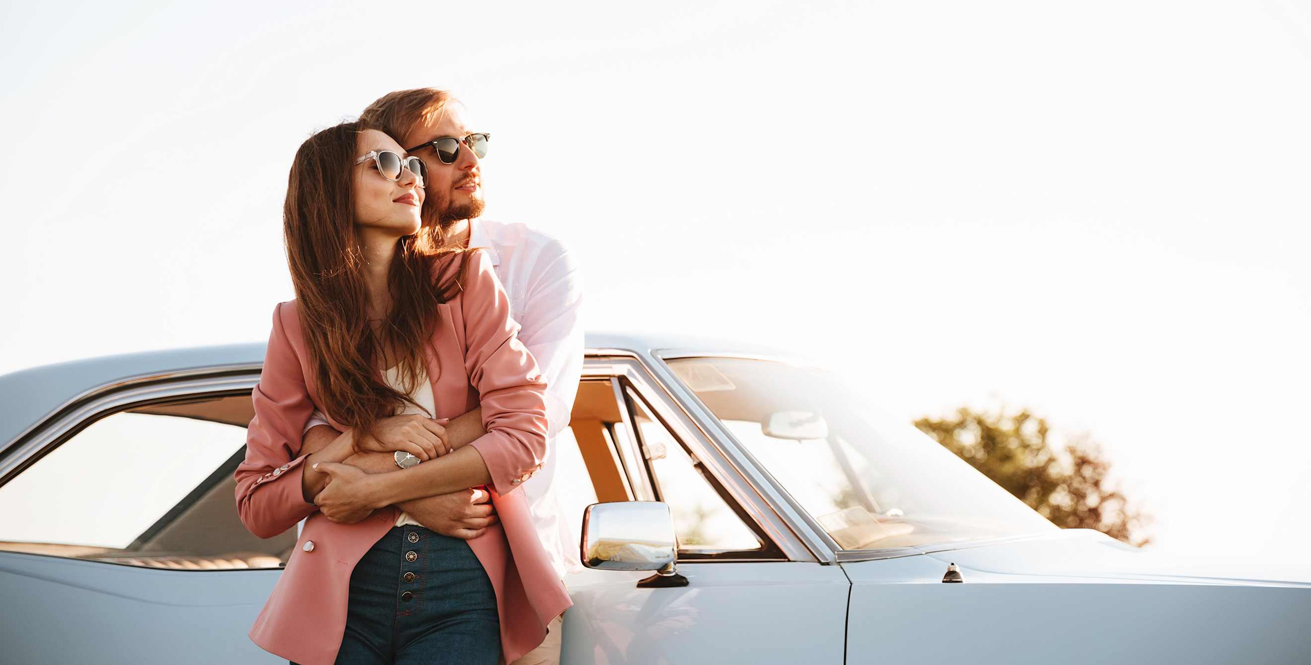 A couple hug in front of a powder blue older American car.
