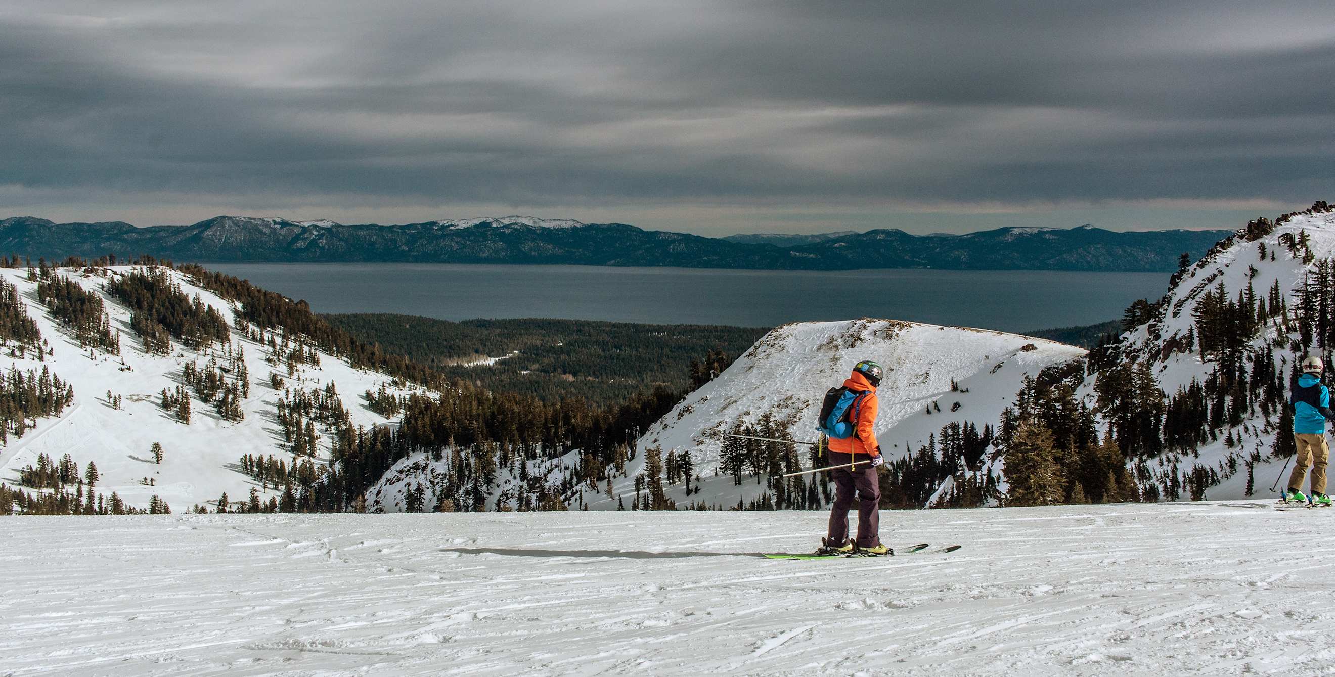 two skiers riding downhill at Alpine Meadows resort with Lake Tahoe in the background