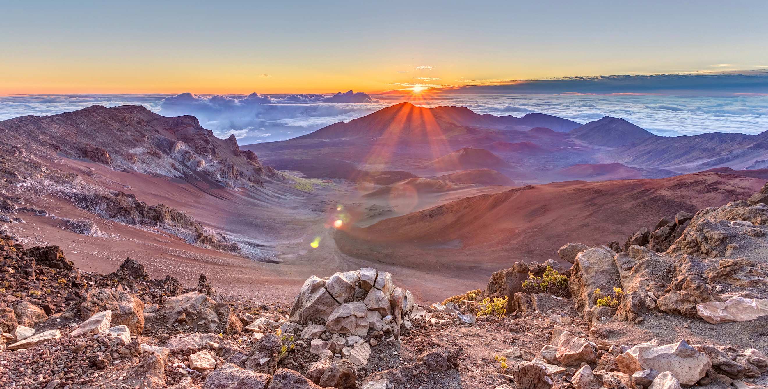 The sun peaks over the clouds into Haleakala at sunrise, image