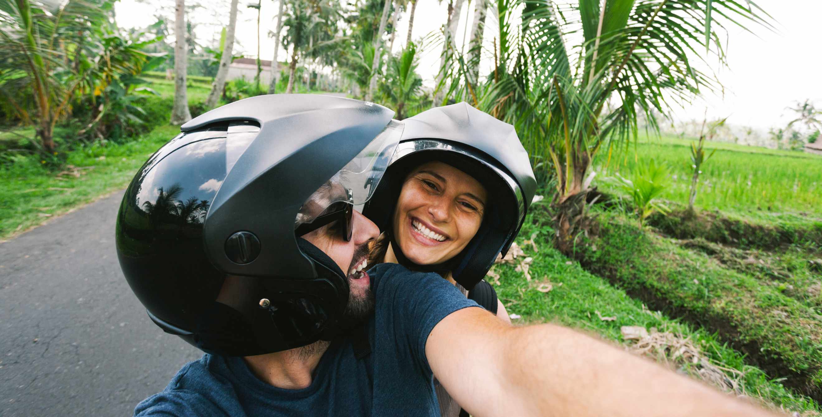 Couple takes a selfie while riding a scooter through a tropical locale