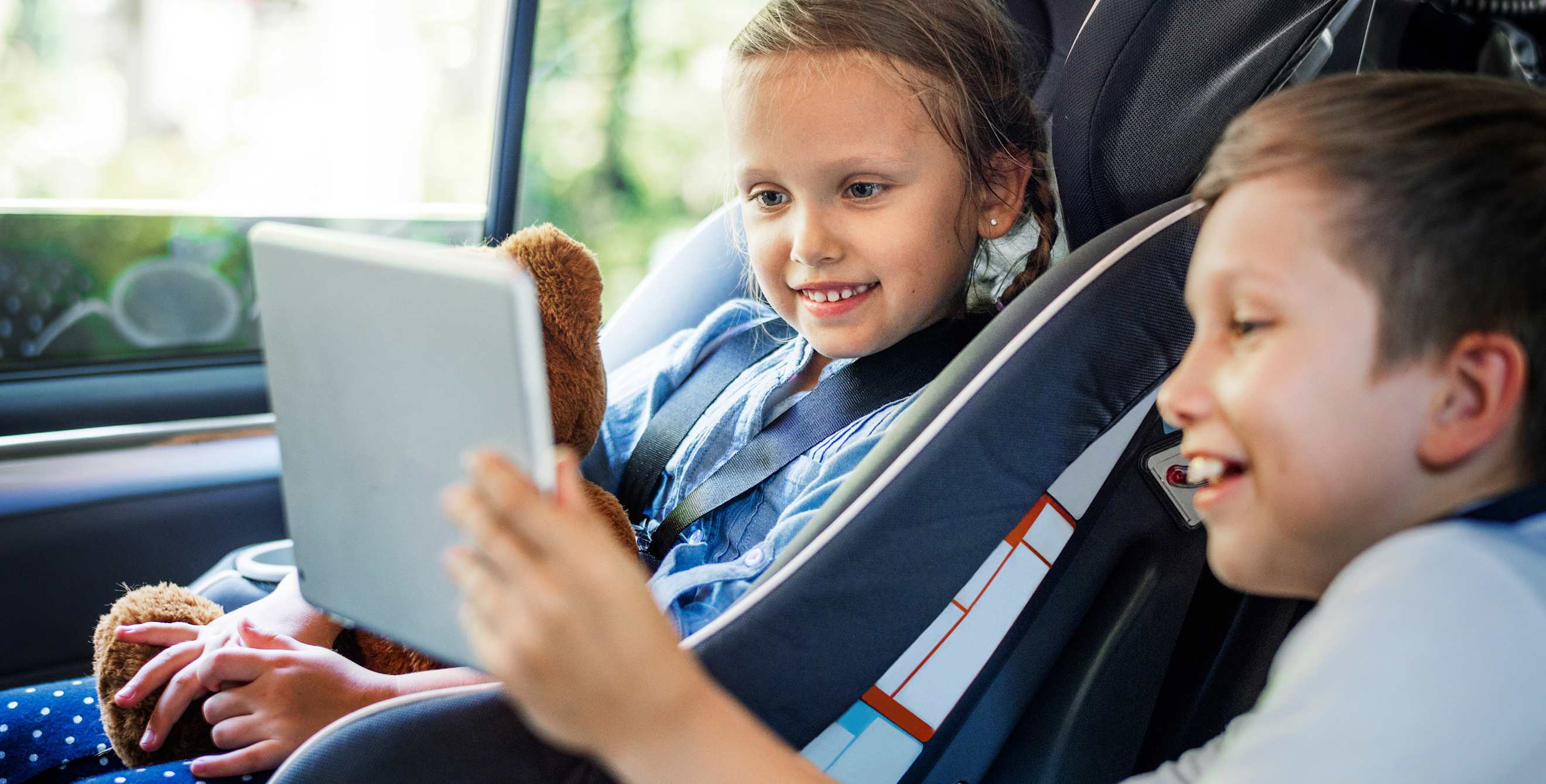 brother and sister look at a tablet in the backseat of a car