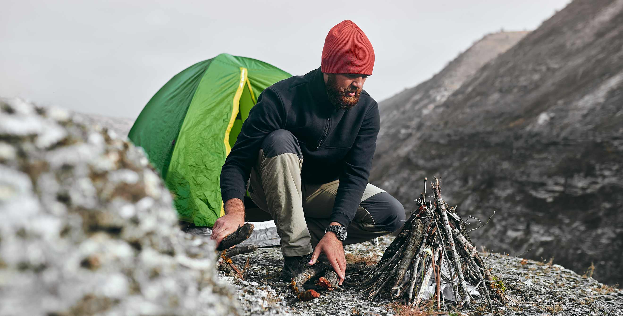 A hiker builds a campfire outside a tent on a mountainside.