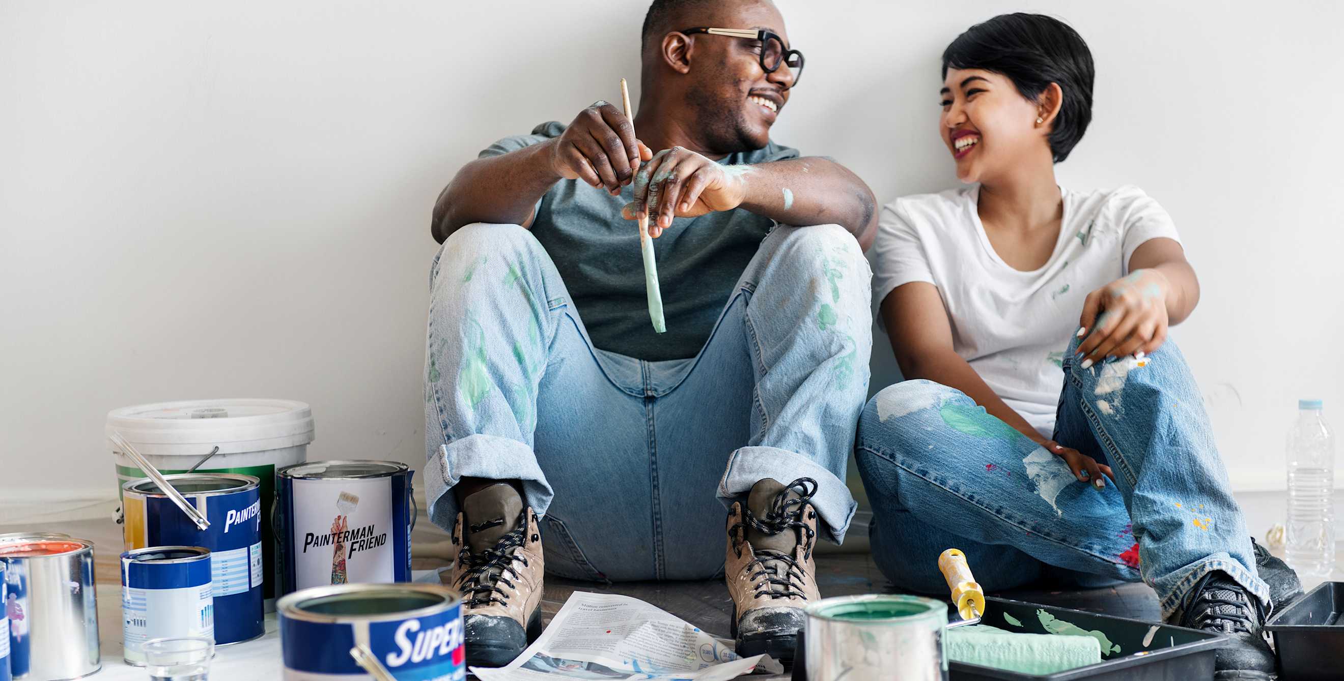 A man and woman sit against a wall before painting
