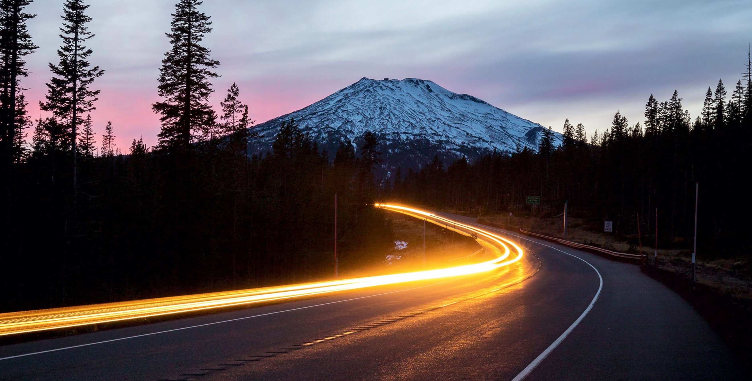 oncoming traffic headlight trails bend along a mountain highway at dusk