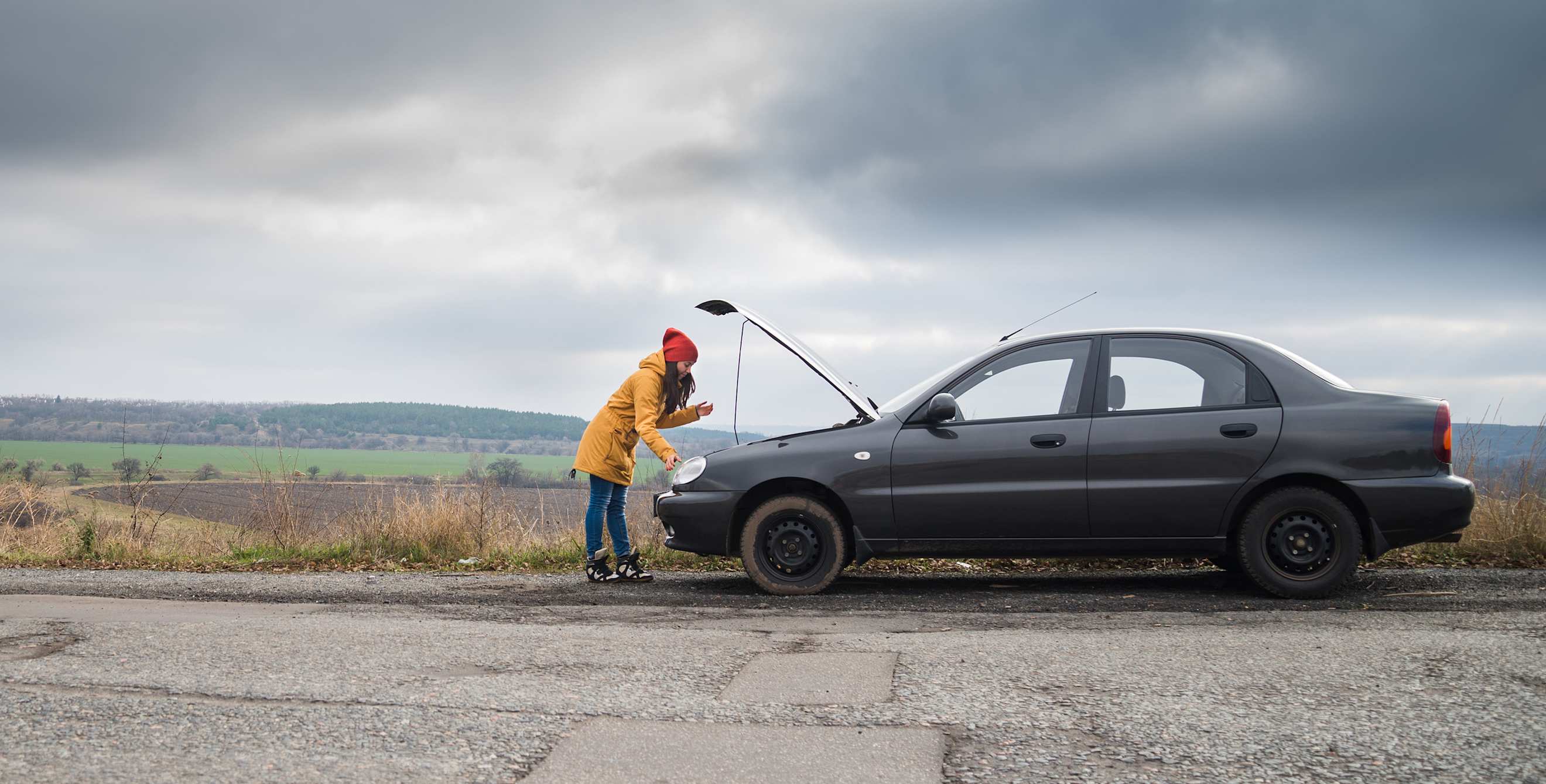 A driver looks under the hood with their car broken down on the side of the road.