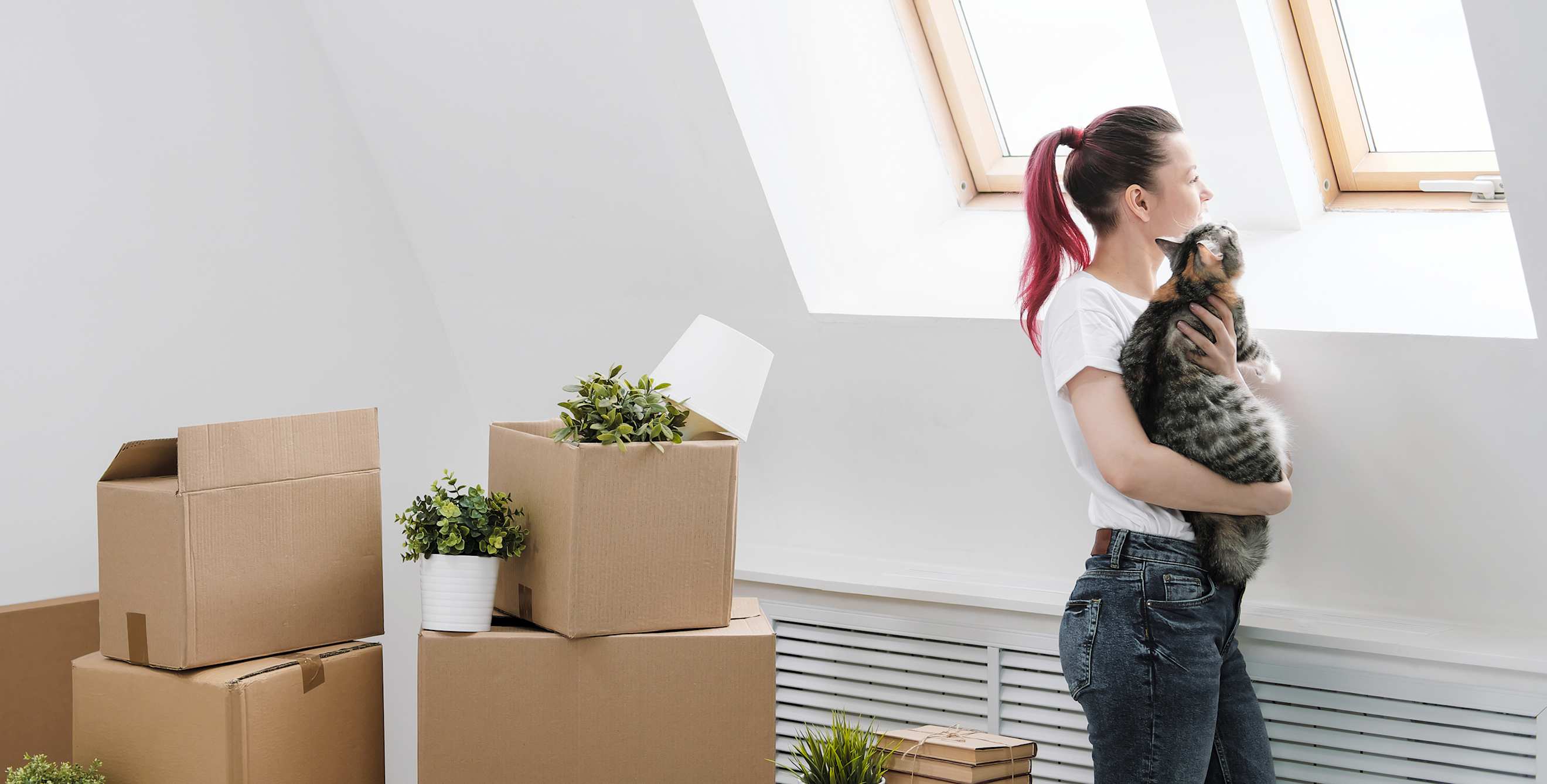 A woman stands in moving boxes with her cat in her arms.