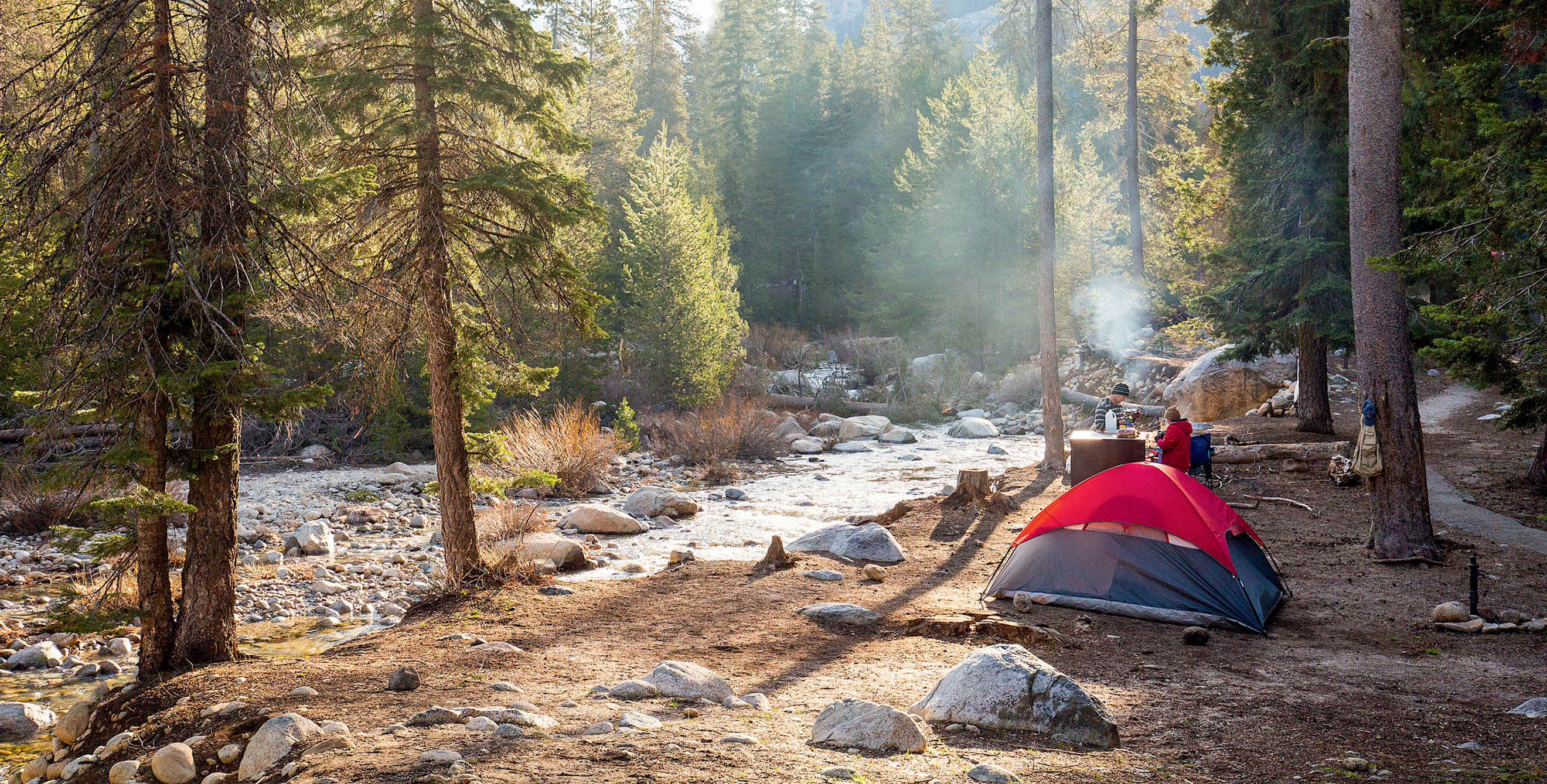 pair of campers with tent pitched beside stream are dressed for cool temps on sunny day at Dorst Creek Campground in Sequoia National Park, California