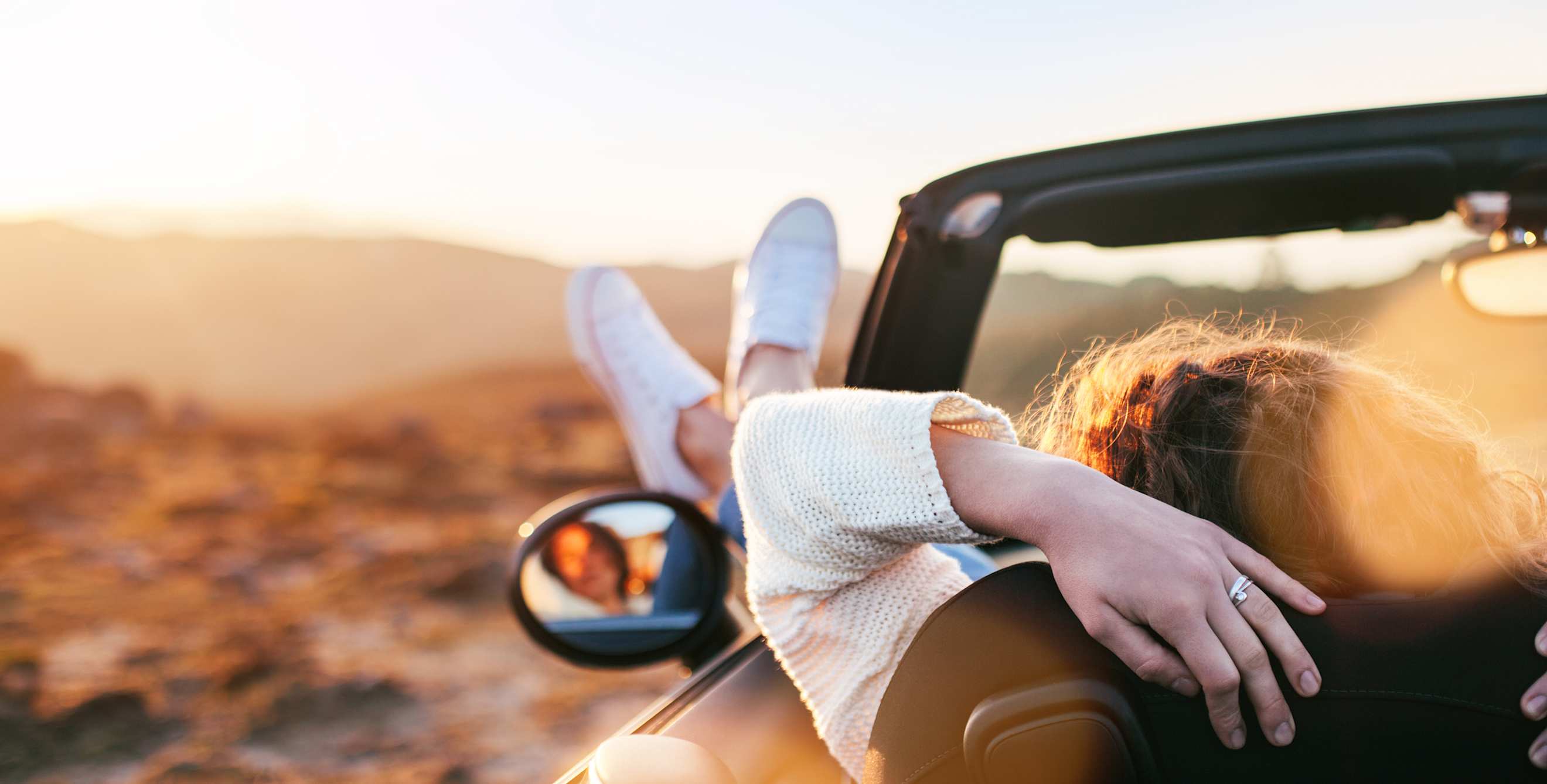 A woman sits with her feet out the window of a convertible at sunset.