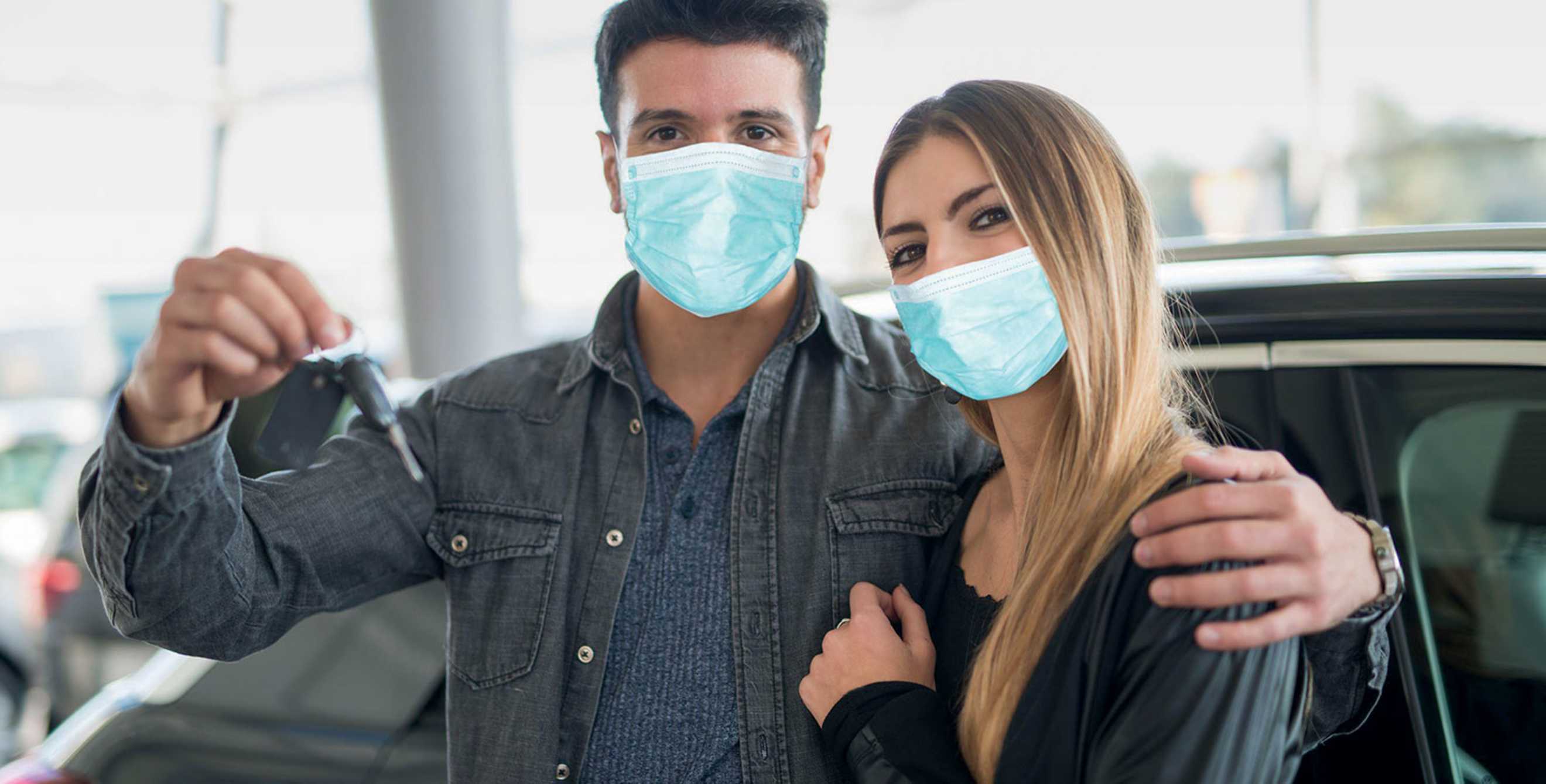 Man with arm around female partner, both wearing disposable protective masks, holds up car keys
