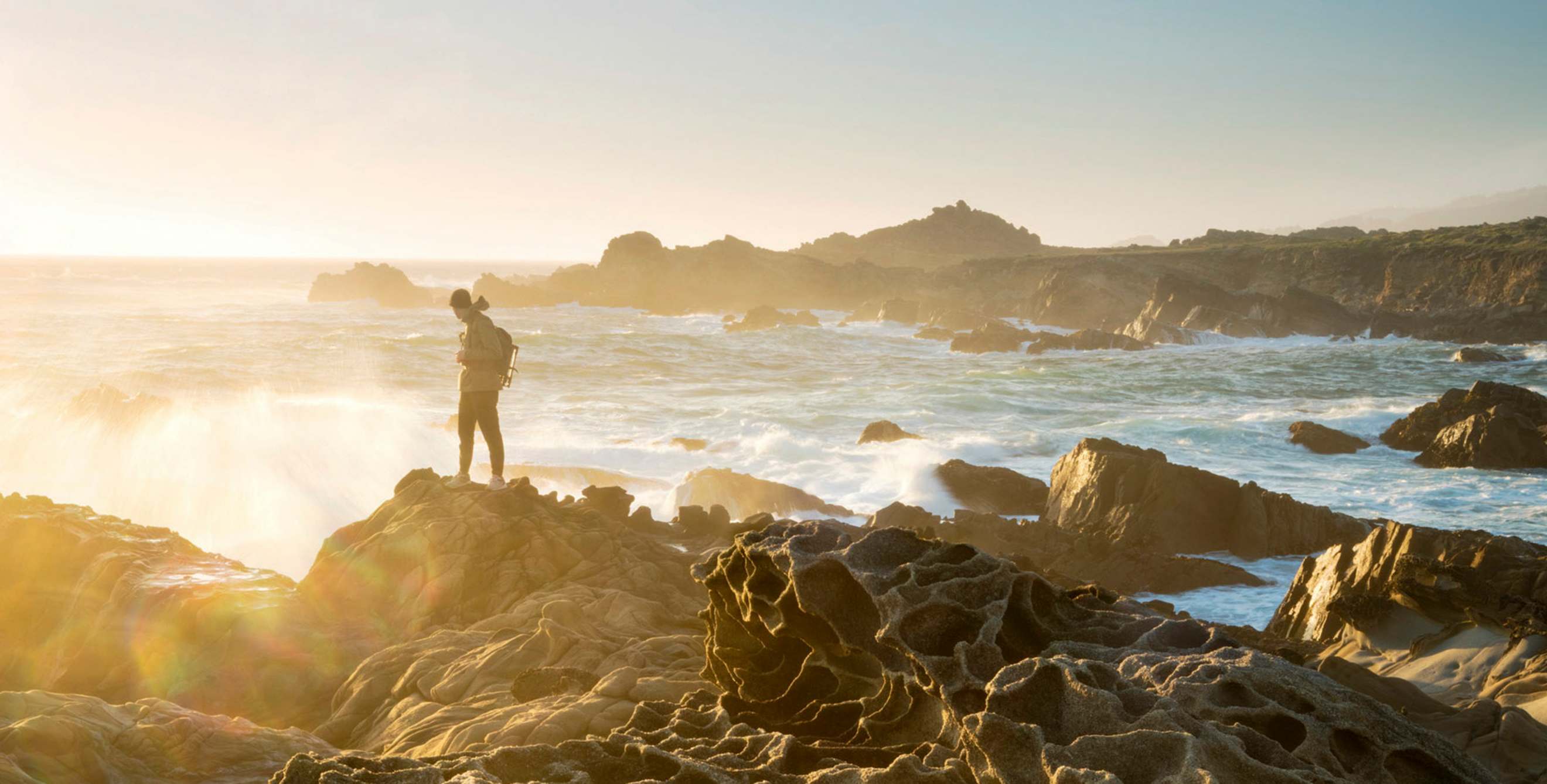 Hiker dressed warmly and standing on the honeycombed rocks known as tafoni looks out to sea at sunset at Salt Point State Park on the Sonoma Coast in California