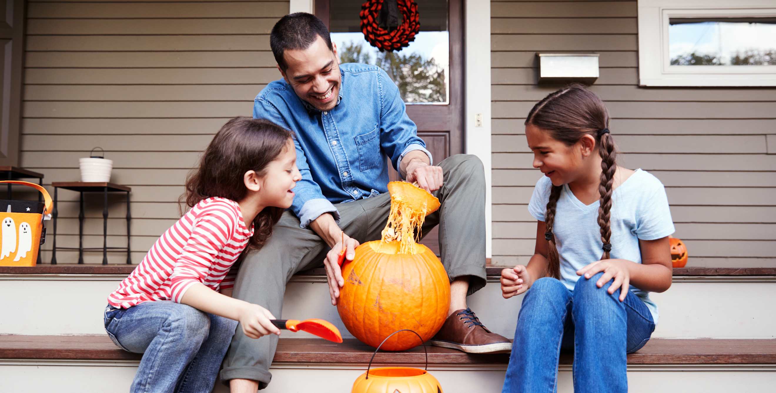 A dad carves a pumpkin with his daughters on their porch.