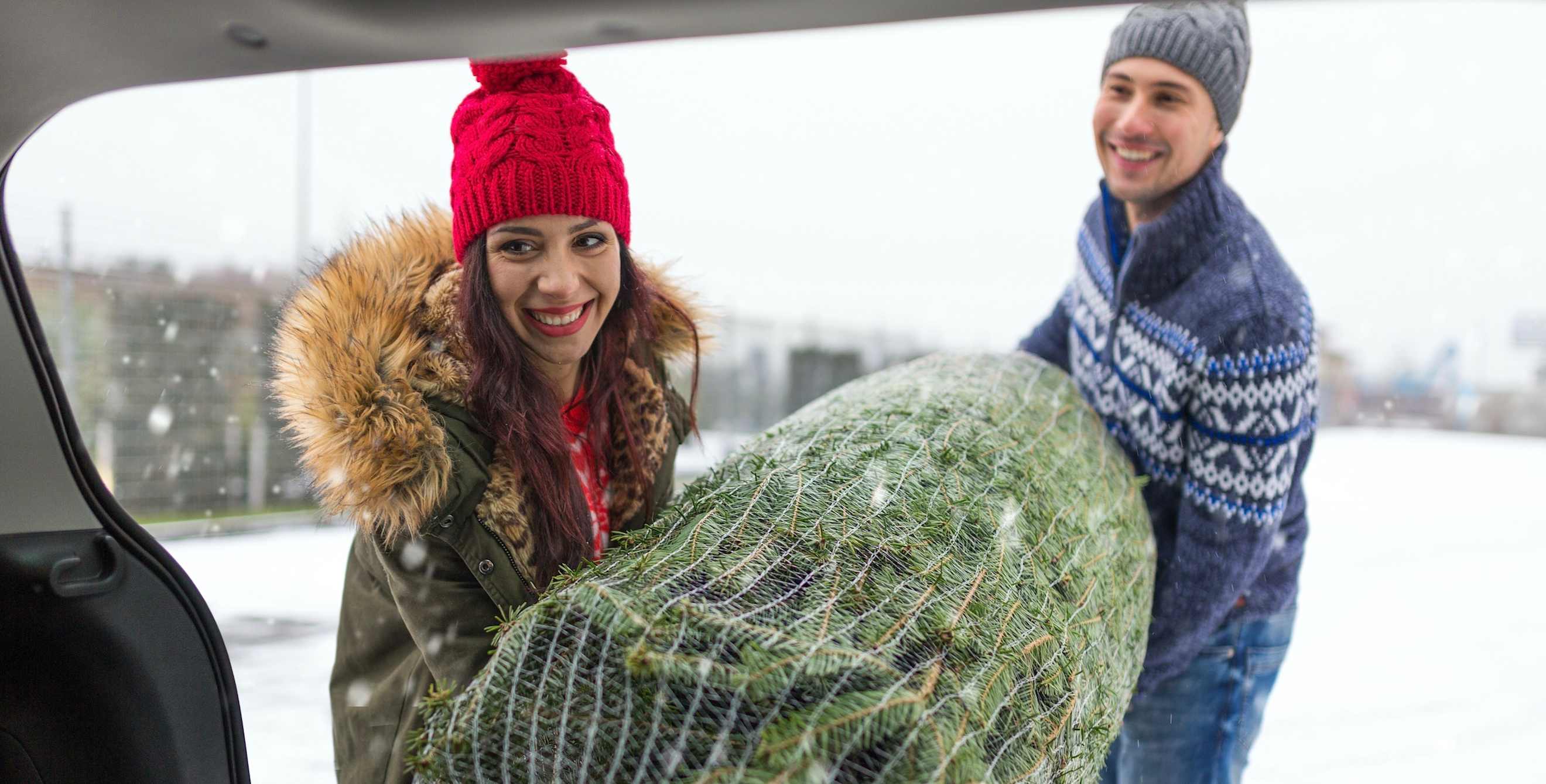 a smiling couple puts their Christmas tree into their hatchback vehicle with snow blowing outside