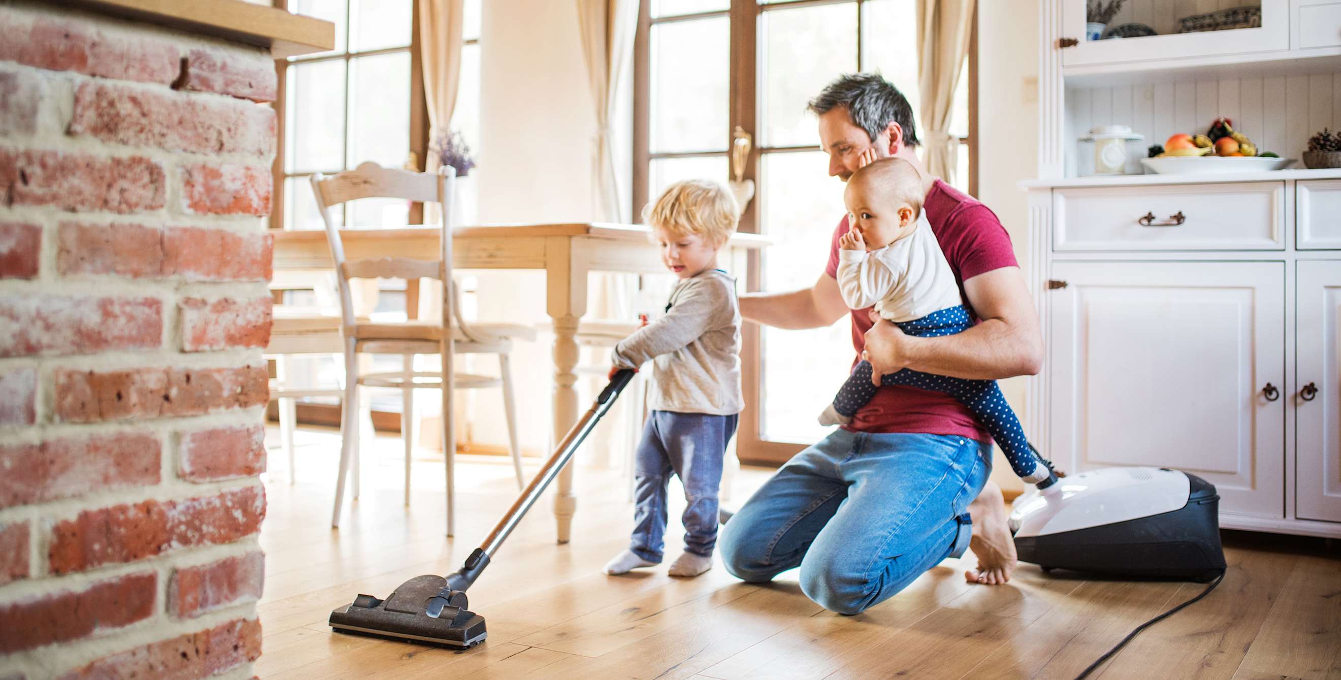 A father vacuums the floor with his child.