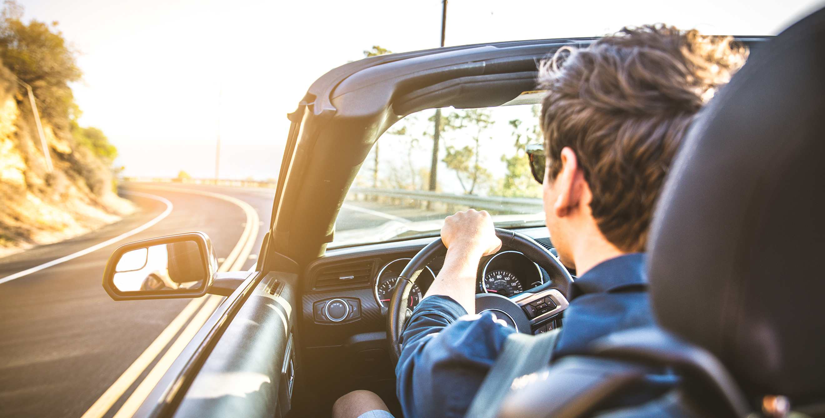 A man drives a convertible on the California coast during golden hour.