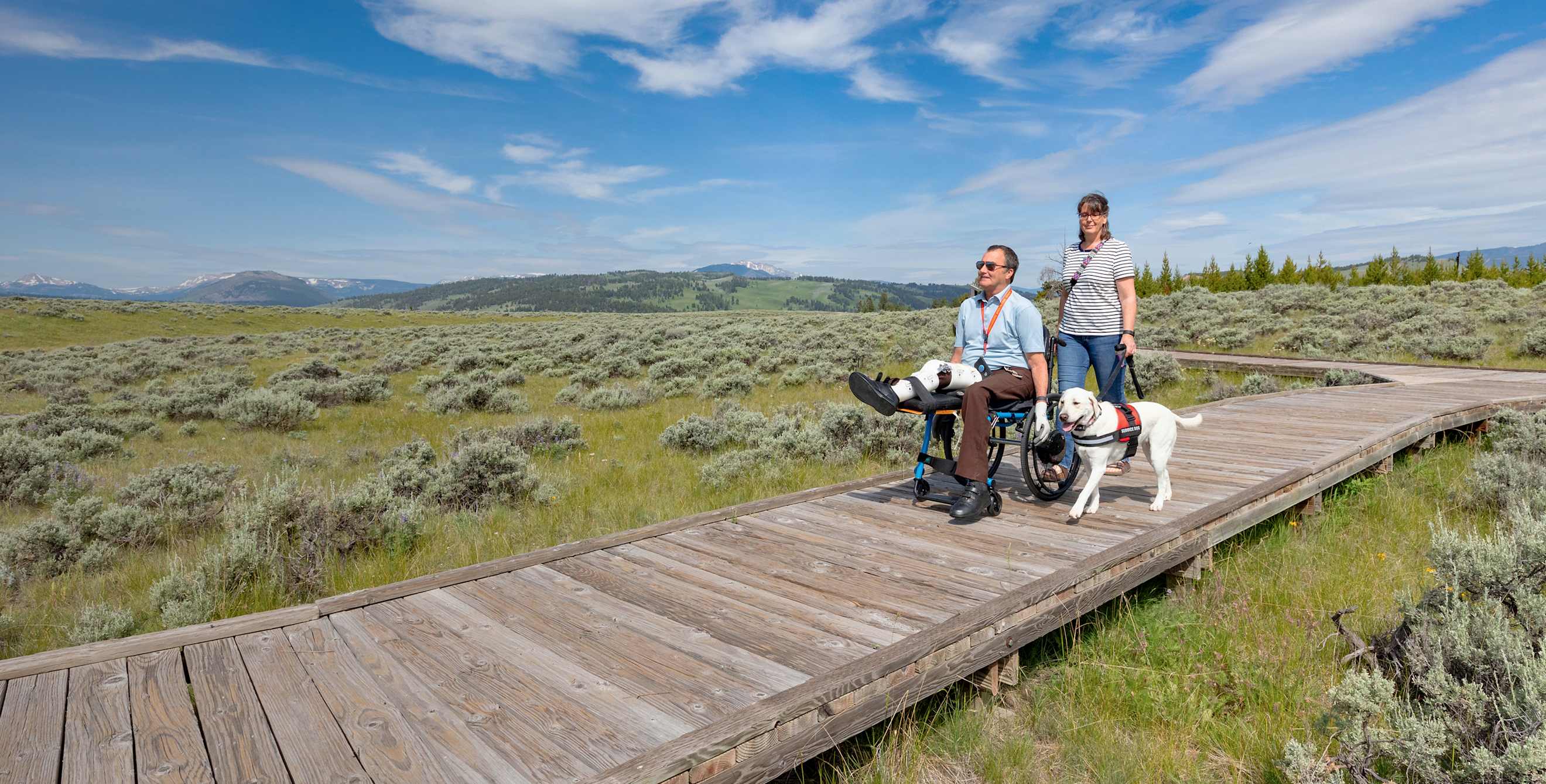 A couple and their service dog hike on a boardwalk trail in Yellowstone National Park.
