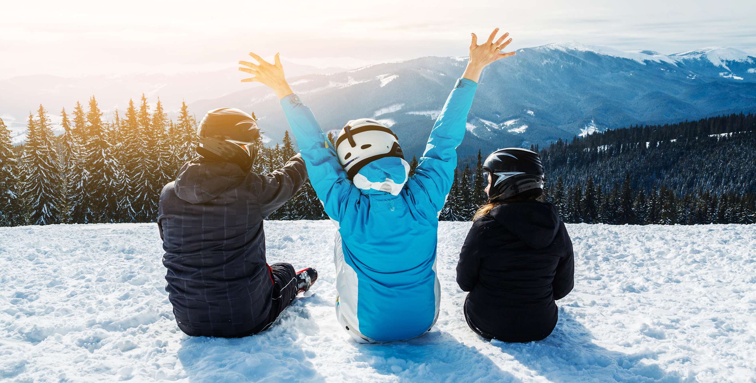 a family sitting on snow overlooking a snow-topped mountain with the sun