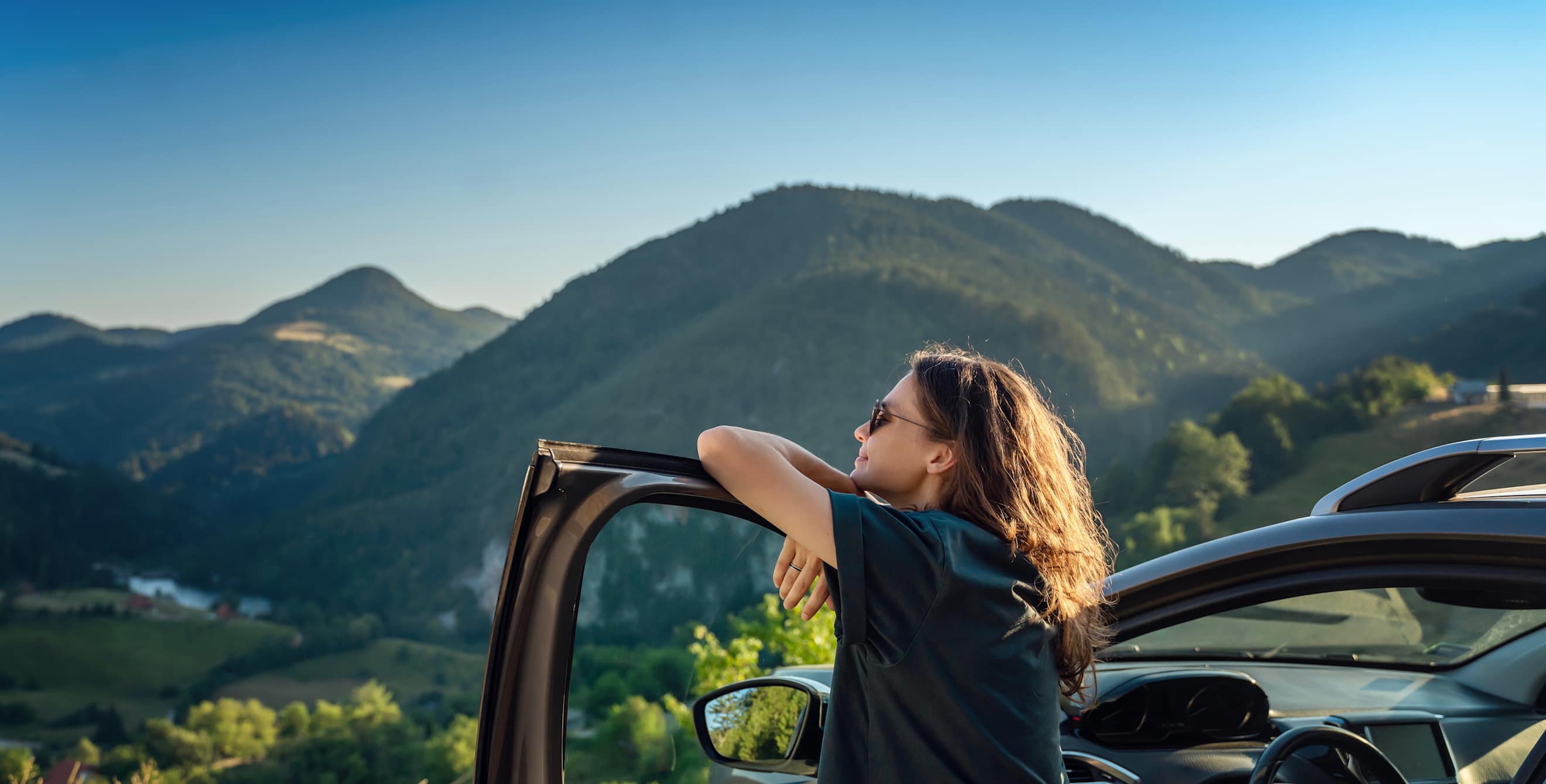 A woman stands by her open car door and looks out a mountain view.
